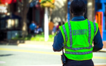 A security guard in a neon green vest with reflective stripes stands outdoors during the day, facing away from the camera. The word 