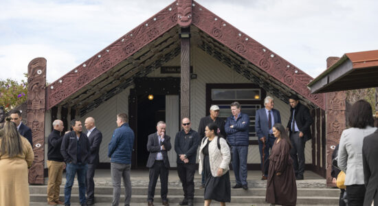 A group of people gathers outside a carved Māori wharenui with intricate red designs. Some individuals are conversing while others observe quietly. Two women walk across the foreground, and the green lawn contrasts with the cloudy sky above.