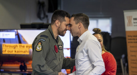 Two men share a hongi, a traditional Māori greeting, during a ceremony. One wears a military uniform with New Zealand Air Force insignia, and the other wears a white shirt. A woman in a red top is visible in the background, with a Ringa Hora banner to the side.