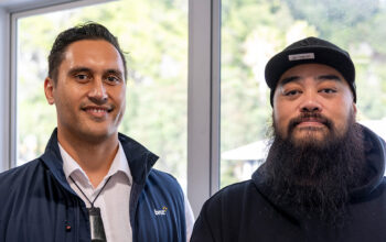 A close up of two people looking towards the camera. One is wearing a blue vest and white shirt. The other is wearing a cap and jumper.