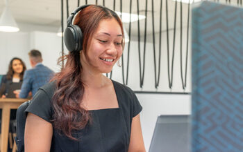 A close up of a person on an angle. They are sitting down with headphones on. It looks like they are in an office environment with other people in the background.