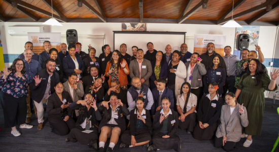 A large group of people posing together inside a meeting room with wooden beams. They are smiling, waving, and making gestures for the photo. Event banners with 'Ringa Hora Workforce Development Council' are visible in the background.