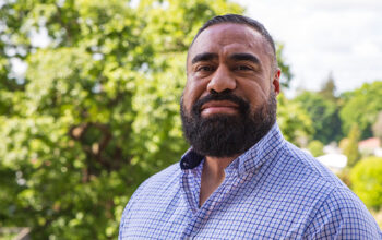 A close up of a person wearing a checkered blue shirt. In the background are bright green leaves from a tall tree.