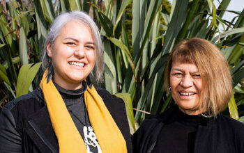 Two wahine Māori (Māori women) standing in front of a flax bush.