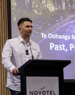 A side angle of a person behind a podium giving a presentation. In the background is screen with a presentation on it.