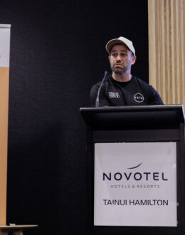 A person standing behind a podium giving a presentation. He is wearing a cap and a black t-shirt. On the side of him is a banner. In the background is a screen.