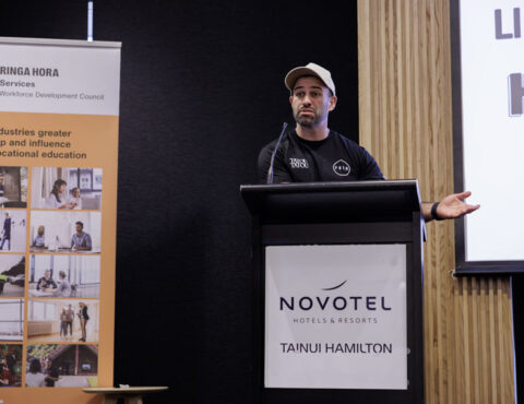 A person standing behind a podium giving a presentation. He is wearing a cap and a black t-shirt. On the side of him is a banner. In the background is a screen.