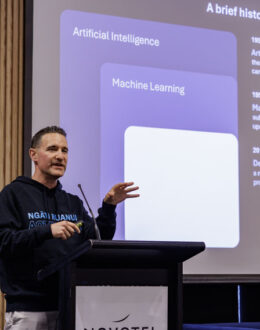 A person standing in front of a podium giving a presentation. In the background is a screen with a presentation on the history of artificial intelligence.