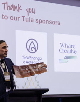 A side angle of a tane Māori (Māori man) doing a presentation. He is wearing a blue suit. In the background is a screen with different business logos.