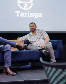 Three people sitting down on blue couches. In the background is a projected image with a sign reading Taringa.