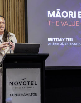 A wahine Māori (Māori women) standing behind a podium and giving a presentation. In the background is screen that shows a slide about Māori Business, and the value of whānau.