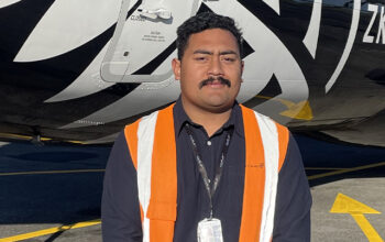 A person standing in front of a black and white plane. They are wearing a blue shirt and a bright orange Hi-Vas.