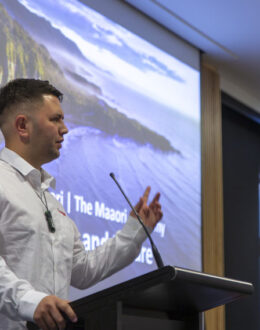 A side angle of a person presenting behind a podium. In the background is a screen wth an ocean and mountain range on it.