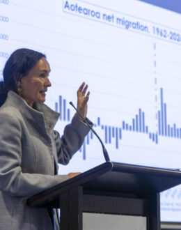 A side angle of a person standing behind a podium and doing a presentation. In the background is a screen that shows a bar graph of Aotearoa's migration rate.