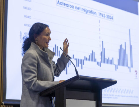 A side angle of a person standing behind a podium and doing a presentation. In the background is a screen that shows a bar graph of Aotearoa's migration rate.