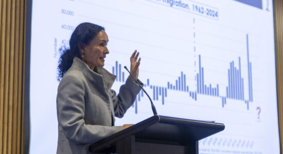 A side angle of a person standing behind a podium and doing a presentation. In the background is a screen that shows a bar graph of Aotearoa's migration rate.