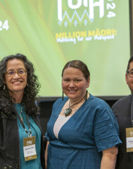 Three people posing together for the camera. In the background is a screen welcoming people to the 2024 Tuia event.