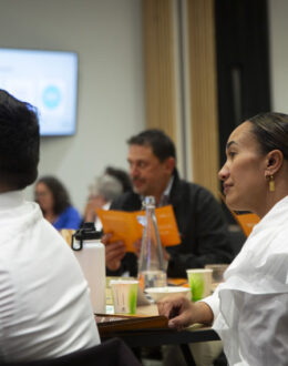 A side angle of a person looking towards the front of the room. In the foreground and background are other people sitting down.