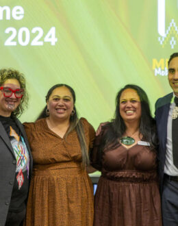 Four people posing together for the camera. They are dressed in formal attire. In the background is a projection image of the Tuia event.