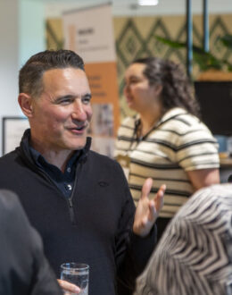 A close up of a person standing with two other people having a conversation. They have a glass in one hand, and their other hand is making a gesture.