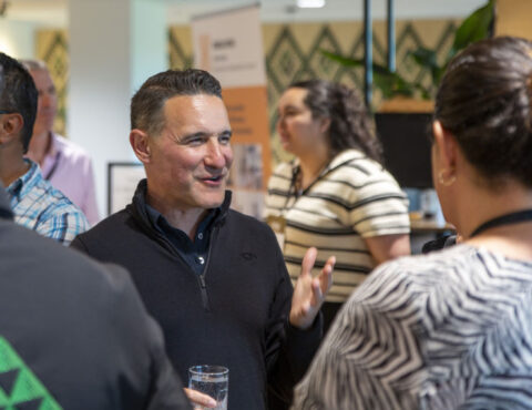 A close up of a person standing with two other people having a conversation. They have a glass in one hand, and their other hand is making a gesture.