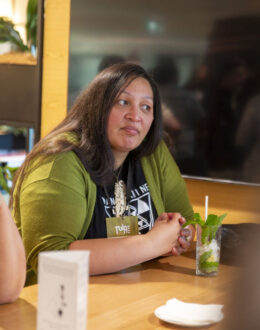 A close up of a wahine Māori (Māori women) sitting at a table. They are staring to the side, and is wearing a green cardigan.