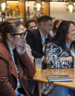 A side angle of two wahine Māori (Māori women) sitting at a table. In the background there are several people standing off to the side of them.