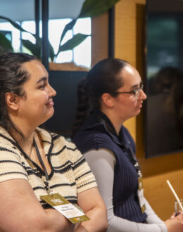 A side angle of two people. In the background is a reflection on. a black TV screen of a room full of people.