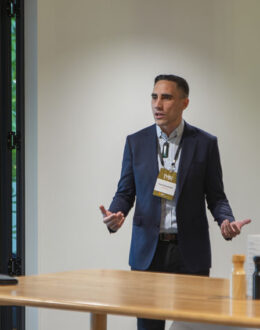 A tane Māori (Māori man) in a room, standing behind a table with their arms gesturing outwards. They are wearing a blue suit, and looks like they are presenting to a group of people.