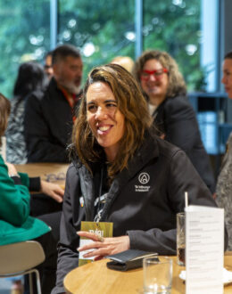 Two wahine Māori (Māori women) having a conversation at a table, in a crowed room of people.