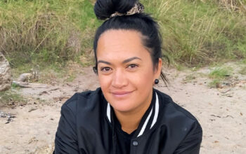 A person with dark hair tied up in a bun, wearing a black jacket with white trim, sitting outdoors near a grassy area.