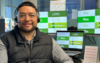 A person with short dark hair, glasses, and a headset, smiling while seated at a desk in a modern office environment. Multiple monitors in the background display green and white graphics.