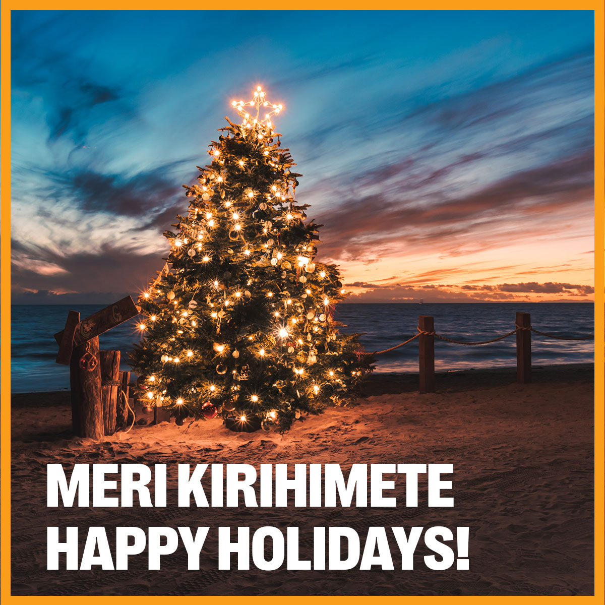 A christmas tree decorated with lights sparkling on a beach at sunset. Text overlay reads Meri Kirihimete Happy Holidays