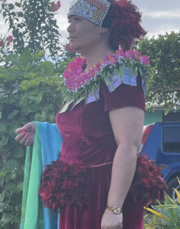 A woman dressed in traditional Tongan attire stands outdoors, adorned with cultural embellishments. She wears a deep red velvet dress with feathered details on the sleeves and hips. Around her shoulders is a floral kahoa (garland) decorated with pink flowers and banknotes. Her headpiece is an intricate, bejeweled design. She holds a multicolored fabric in her left hand while gazing into the distance, with lush green foliage and a parked car in the background.