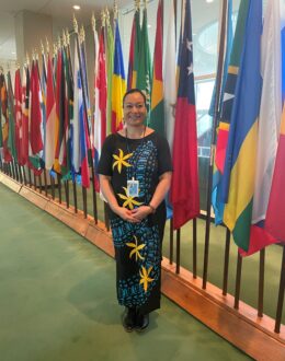 A woman stands smiling in front of a row of international flags inside the United Nations headquarters. She wears a traditional Pacific Island dress in black and blue, adorned with bright yellow floral patterns. Her ID badge hangs from a lanyard, signifying her participation in an official event. The setting features a green carpet and a backdrop of flags representing global unity and diplomacy.