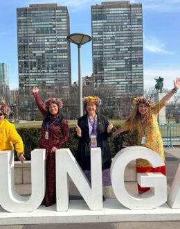 Four women stand in front of large white letters spelling '#UNGA' outside the United Nations headquarters in New York City. They are dressed in vibrant, traditional attire, each wearing a colorful floral headpiece. They pose energetically with arms raised, smiling against a backdrop of modern skyscrapers and a clear blue sky, symbolizing cultural representation and participation at the UN General Assembly