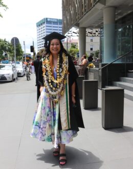 A young woman in a black graduation cap and gown walks confidently outside a university building, smiling. She wears a colorful floral dress underneath and is adorned with multiple traditional Tongan kahoa (garlands) made of shells and gold decorations. The background features a busy city street with pedestrians, cars, and a university sign, indicating a graduation ceremony setting.