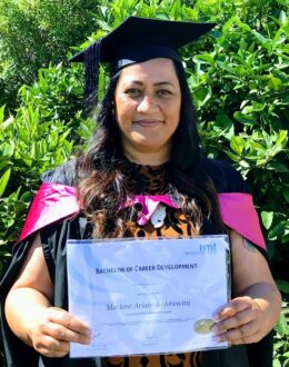 A woman in a black graduation cap and gown stands outdoors in front of lush green foliage. She smiles as she holds a Bachelor of Career Development degree certificate from NMIT. Her gown features a pink stole draped over her shoulders, and she wears a black-and-orange patterned dress underneath.