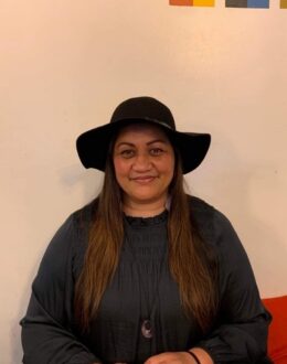 A woman sits in front of a simple beige wall, smiling gently at the camera. She wears a black, wide-brimmed hat and a long-sleeved black dress with pleated detailing. Her long brown hair falls over her shoulders, and she wears a pendant necklace.