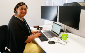 A woman sits at a modern office desk, typing on a laptop connected to multiple monitors. She wears a black blouse and yellow pants, with a blue ID lanyard around her neck. A green reusable coffee cup sits on the desk. She smiles warmly at the camera, with a professional and organized workspace behind her.