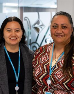 Two women stand side by side, smiling in an office environment. Both wear blue lanyards with ID badges. The younger woman on the left has long, curly black hair and wears a dark grey coat over a black top. The woman on the right wears a red and brown geometric-patterned top with long, dark brown hair tied back. Behind them, a glass door features a Māori-inspired design.