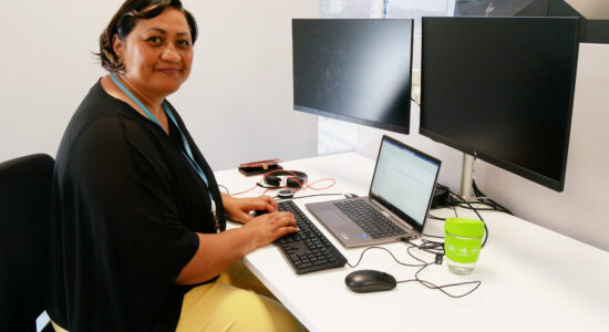 A woman sits at a modern office desk, typing on a laptop connected to multiple monitors. She wears a black blouse and yellow pants, with a blue ID lanyard around her neck. A green reusable coffee cup sits on the desk. She smiles warmly at the camera, with a professional and organized workspace behind her.