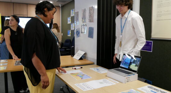 A woman wearing a black top and yellow pants leans over a table, looking at an educational display about water management. A young man with curly hair, wearing a white shirt and a blue ID lanyard, stands behind the table explaining the materials. Various maps, a laptop, and printed resources are spread across the table, with other attendees in the background engaged in discussions.