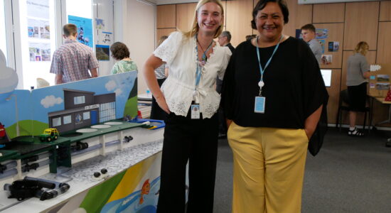 Two women stand in front of a display table showcasing an educational model of water infrastructure. Both wear blue ID lanyards. The woman on the left wears a white blouse with lace details and black pants, while the woman on the right wears a black top and yellow trousers. They smile warmly in a setting with other attendees engaging in discussions and exhibits in the background.