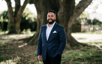 A man with a beard, wearing a navy-blue suit with a white pocket square, stands outdoors under a large tree. He smiles warmly, with a natural setting of grass and sunlight filtering through the branches in the background.