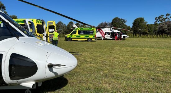 A medical emergency response scene in an open grassy field, featuring two white rescue helicopters and multiple yellow and green ambulances. A paramedic in a high-visibility vest stands near an ambulance, while other emergency personnel attend to a patient near one of the helicopters. The clear blue sky and surrounding trees provide a serene contrast to the urgency of the situation.