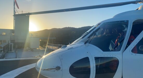 A white rescue helicopter is parked on a rooftop helipad with the sun rising behind distant hills. The cockpit reflects the golden light, and a crew member inside the helicopter appears focused on a task. A windsock and a small rooftop structure are visible in the background, adding to the early morning atmosphere.