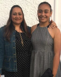 A woman and a young girl stand together, smiling in front of a white textured wall. The woman on the left wears a black polka-dotted dress with a denim jacket, while the girl wears a black-and-white polka-dotted sleeveless dress. The woman’s arm is wrapped around the girl's shoulder in a warm, affectionate pose.