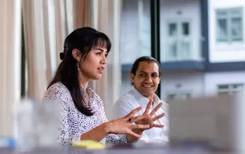 A woman with long dark hair gestures as she speaks during a discussion. She is wearing a white blouse with black polka dots. A smiling man with a white shirt listens attentively in the background, with large windows showing a modern building exterior.