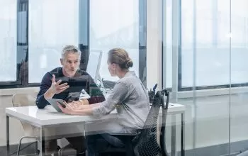 Two people sitting in an office. There is a large glass door in front of them.